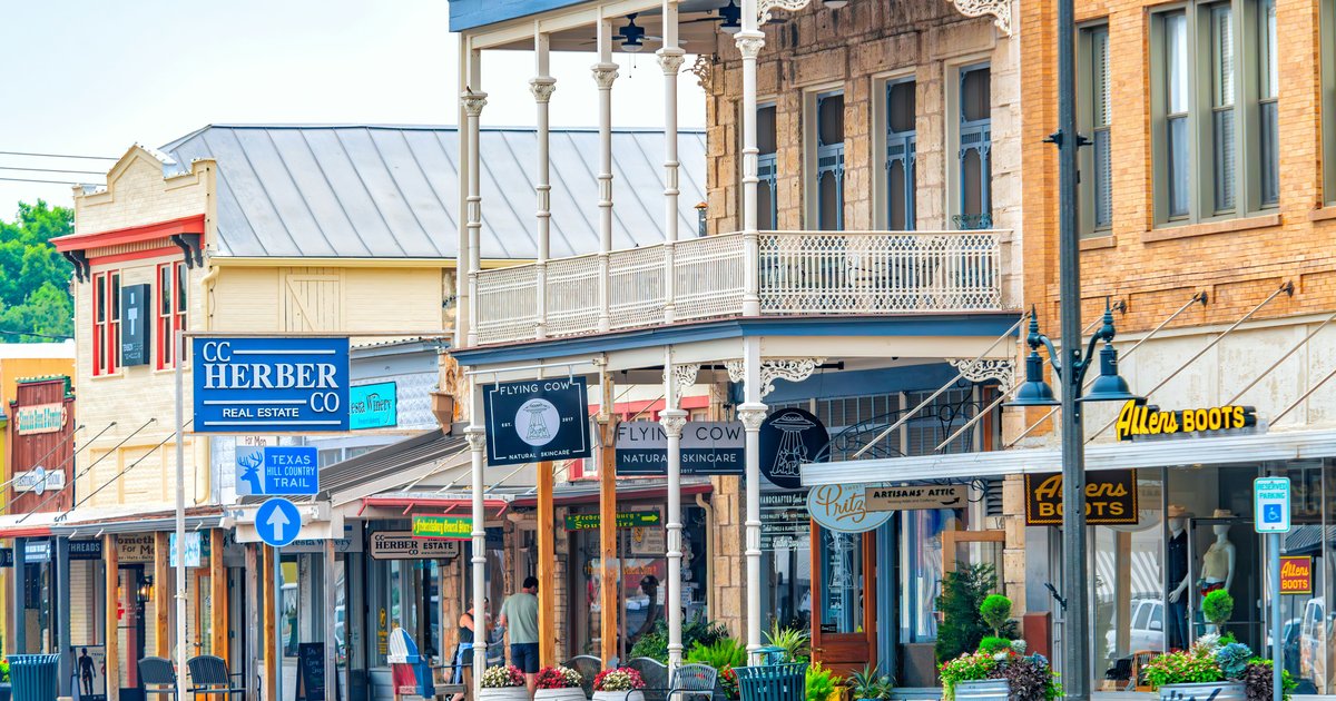 Historic two-story storefronts with wraparound balconies line a small-town Main Street, with hanging flower baskets and awnings over ground-floor shops