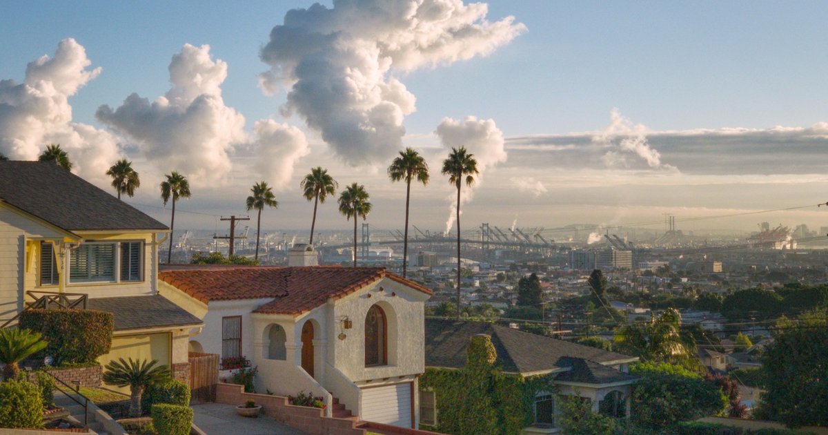 A 1920s Spanish Colonial Revival house with a clay tile roof and arched entry beside an older wood-frame home on a palm-lined Los Angeles street