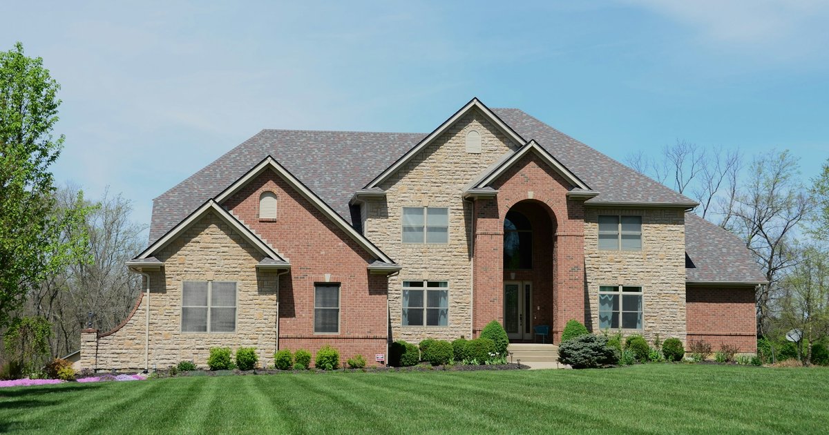 Two-story brick and stone house with arched entry and a wide manicured lawn, illustrating the established single-family housing style found in Far North Dallas
