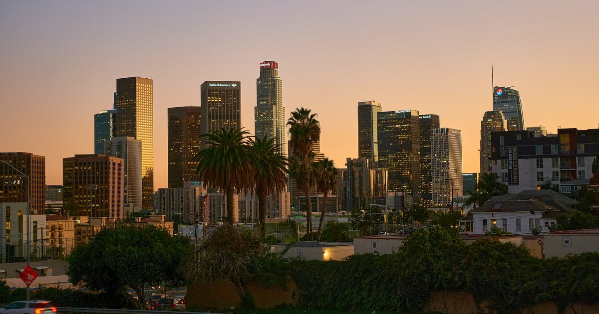 Tall Los Angeles residential and office towers rising behind a row of palm trees along a wide boulevard