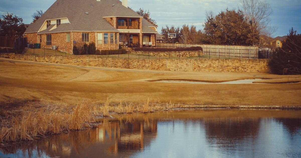 Two-story brick home with a covered porch and stone retaining wall beside a pond in North Texas