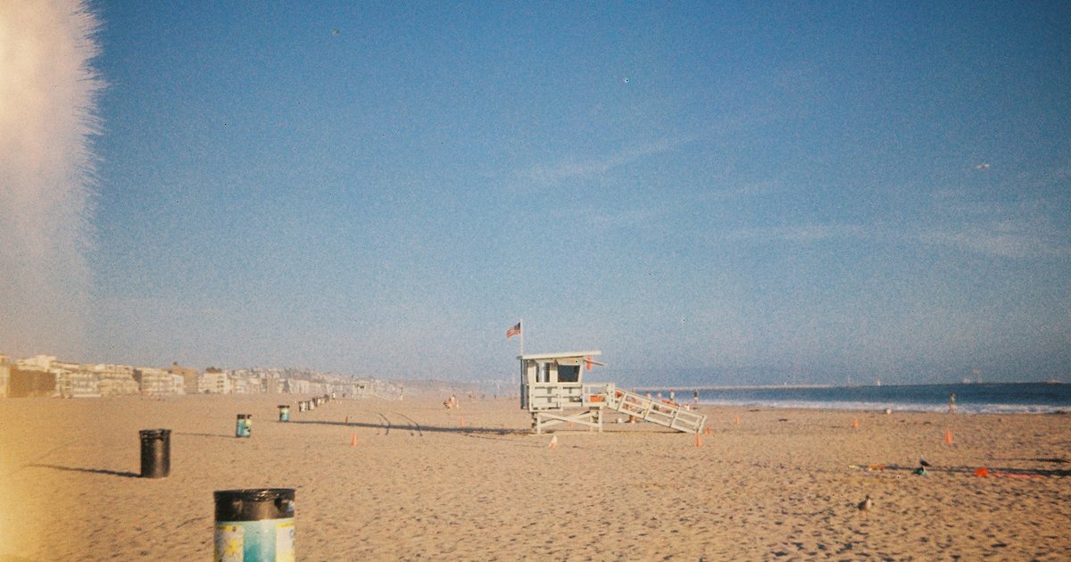 Lifeguard tower on a wide sandy beach with a row of beachfront buildings in the distance under a clear blue sky