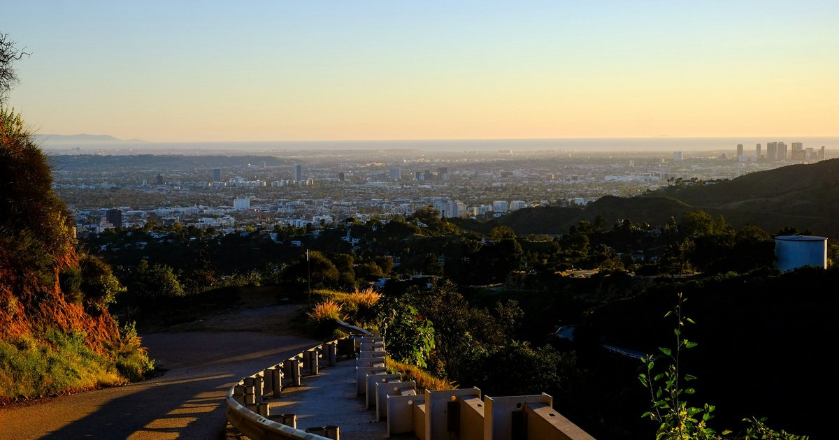 A canyon road curving along a hillside above the Los Angeles basin at sunset, with the Westside skyline visible in the distance