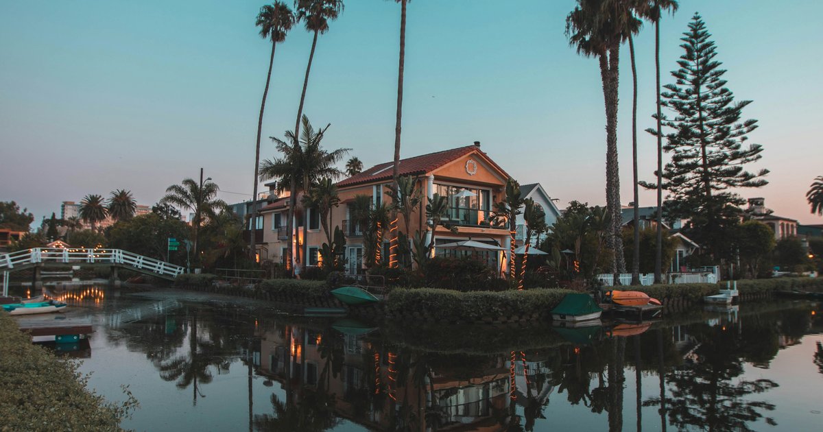 Venice Canals at dusk with a white pedestrian footbridge, moored dinghies along the seawall, and canal-front homes reflected in the water