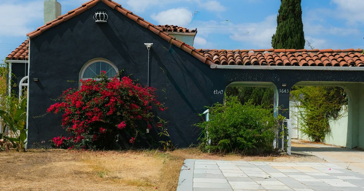 Dark stucco Spanish-style house with a clay tile roof and a blooming bougainvillea beside a paved driveway
