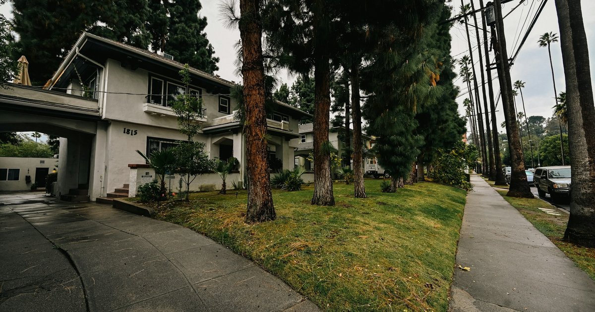 Stucco two-story house with a wide carport arch and a lawn lined with tall conifers and palms along a Los Angeles street